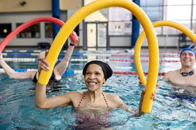 Pessoas fazem hidroginástica na piscina; em primeiro plano há uma mulher parda sorrindo e ao fundo há um homem branco também sorrindo