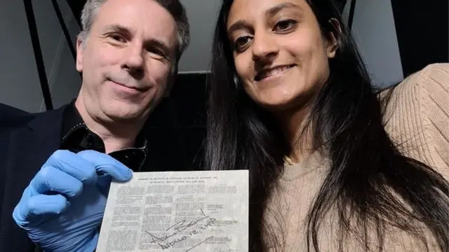 Spencer e Dhara posam para uma selfie com um documento legal que foi à lua.