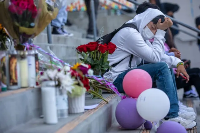 Jovem sentado em escadaria observa homenagens para Melanie ali colocadas, como velas e flores