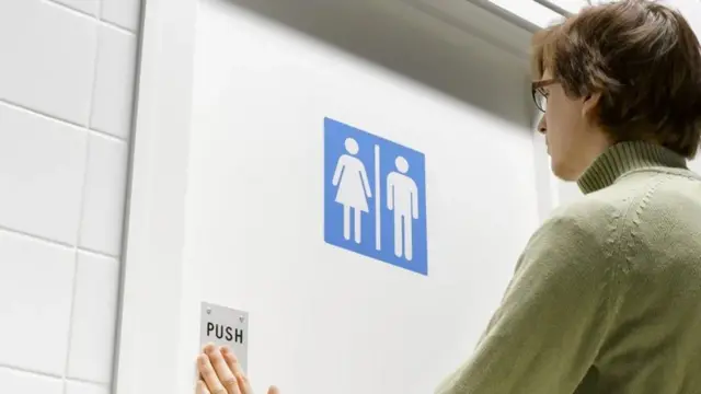 Uma pessoa com a mão na porta de um banheiro cuja placa indica que é unissex