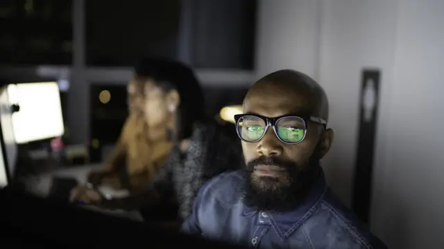 Homem e mulheres no pano de fundo com rostos iluminados por telas de computador em escritório à noite