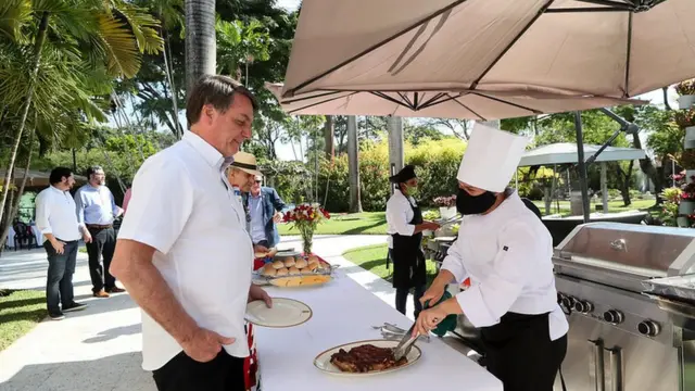 Bolsonaro em frente a mesa em que churrasqueira serve carne, em área aberta e arborizada