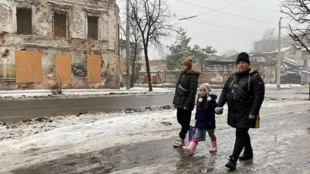 A família caminha para a escola passando por vidros quebrados e edifícios destruídos