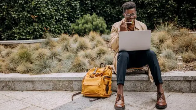 Homem sentado no meio fio, comendo e sorrindo em direção a laptop