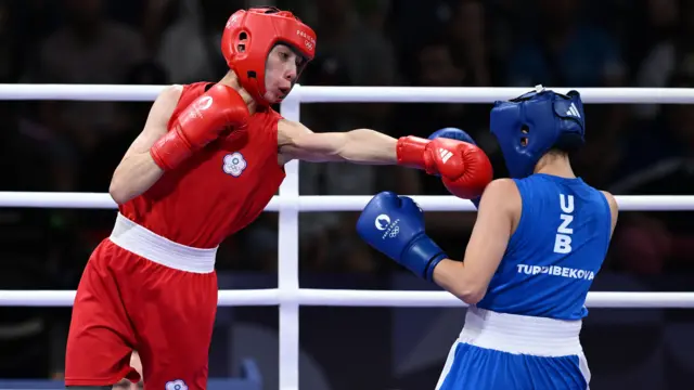 De um lado, uma lutadora de boxe, com luvas e proteção na cabeça vermelhos, do lado direito, de costas, outra lutadora, vestindo tudo azul, recebe um golpe. Ambas estão em um ringue