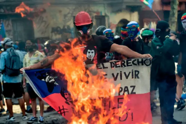 Manifestante com máscara e segurando bandeira do Chile queimando 