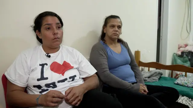 Pamela e Michele sentadas em cama, observando algo na frente delas com olhar sério