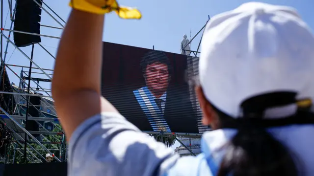 Foto colorida mostra apoiador(a) de Milei com camiseta da argentina, de costas, em frente a um telão com foto de Milei tomando posse