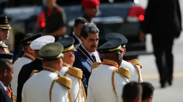 Maduro, um homem branco, de cabelos grisalhos e bigode, está no meio de vários homens vestidos com uma roupa formal branca e boina