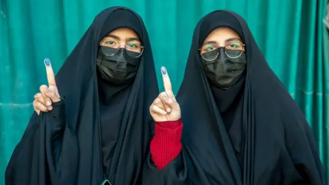 Duas mulheres com rosto e corpo coberto por trajes típicos mostram dedo indicador manchado com tinta 