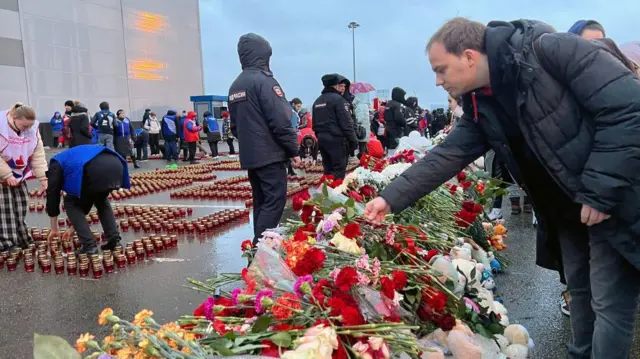 Pessoas colocando flores no chão e acendendo velas em homenagem às vítimas