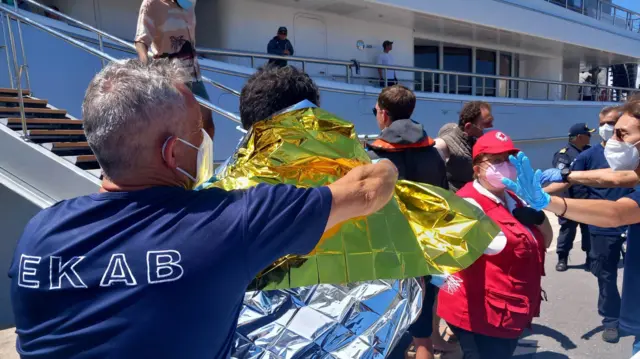 Agente com máscara coloca manta térmica sobre homem, que aparece de costas e perto de grande barco