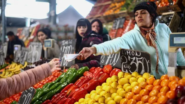 Vendedora em frente a banca de frutas e legumes entrega troco a cliente