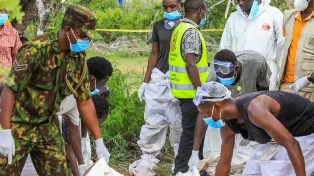 Homens negros, um com uniforme militar e outro com roupas anticontaminação, retiram corpo (não visível na foto) de uma área cheia de policiais e peritos; ao fundo uma faixa da polícia impede o acesso ao local, em um campo gramado