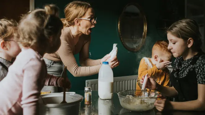 Família de pessoas brancas e loiras na cozinha, a mãe segura algo nas mãos e a criança pequena chora 