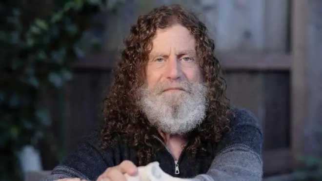 Foto colorida do filósofo Robert Sapolsky, um homem branco de olhos claros, cabelo cacheado na altura do pescoço e barba branca, vestindo uma camiseta azul de lã