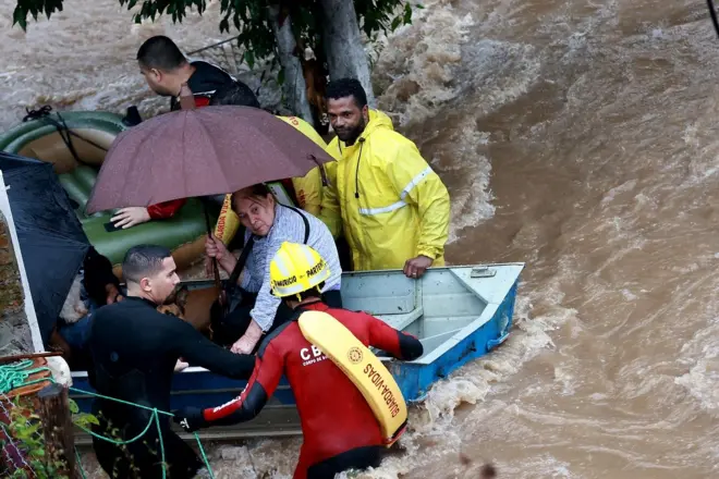 Bombeiros e outras pessoas resgatam uma mulher idosa em um barco em uma região alagada