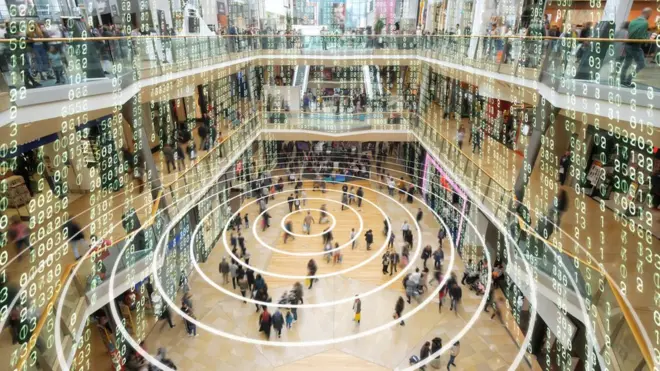 Fotografia colorida mostra pessoas em um shopping e números brancos tipo matriz flutuando em todo o ambiente