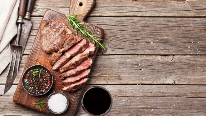 Carne grelhada e fatiada em uma tábua, cercada por potinho de sal, primenta e com um ramo de alecrim ao lado