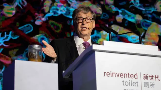 Bill Gates aponta para um pote com cocô, em um evento