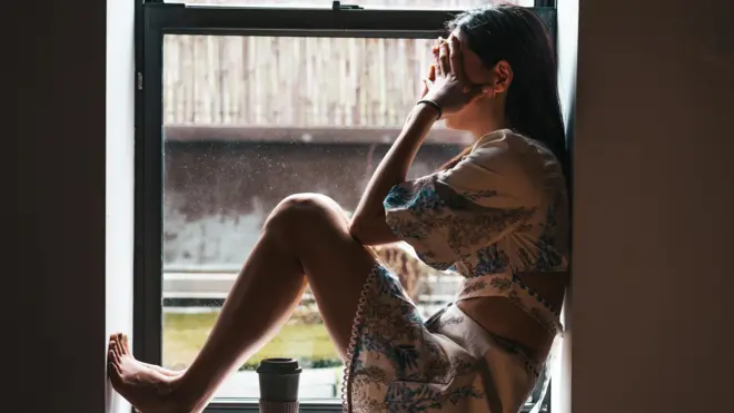 Uma mulher sozinha sentada em frente a uma janela com as mãos no rosto