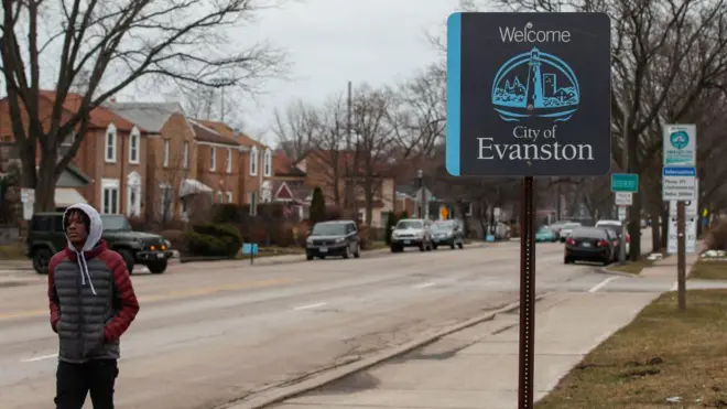 Homem caminhando na rua, passando por placa em que se lê: Bem-vindo à cidade de Evanston em 2021