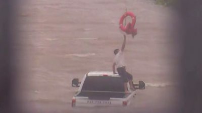 A man sits on top of a half-submerged pickup truck and reaches for a life-ring dangling from above