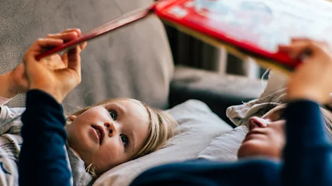 A child laying in bed next to her parent reading a book (Credit: Getty Images)