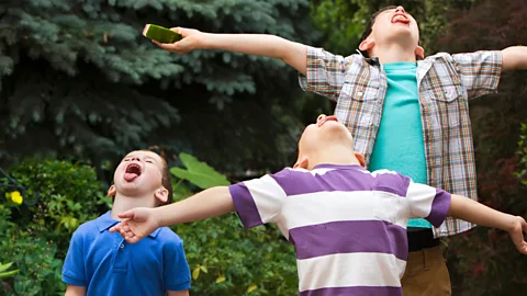 Three boys with tongues out/ heads up to taste the rain, green bushes in background (Credit: Getty Images)