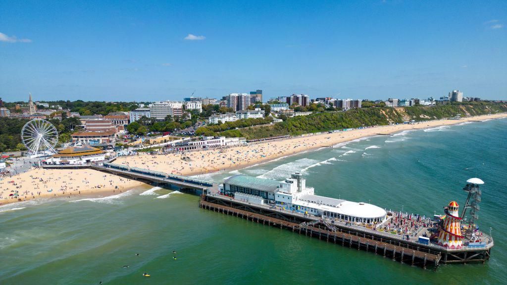 An aerial view of Bournemouth Pier and beach is seen. It is a sunny day and the beach is busy.