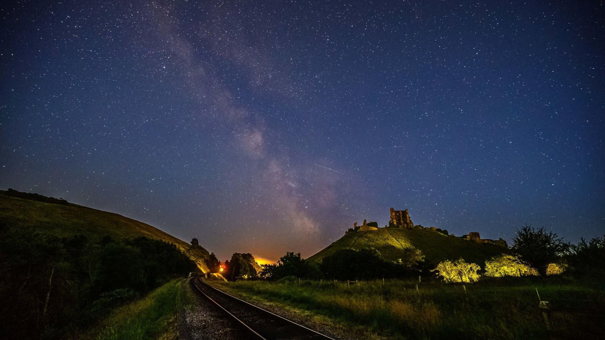 FRIDAY - The night sky over the Dorset countryside. A railway line runs through the centre, with the ruins of Corfe Castle visible on a hill to the right with a green hill to the right of the tracks It is night so the colours are muted, but in the distance you can see the glow from the street lights in Corfe Castle village. In the dark sky above there are thousands of stars with the milky way running through the centre of the sky.