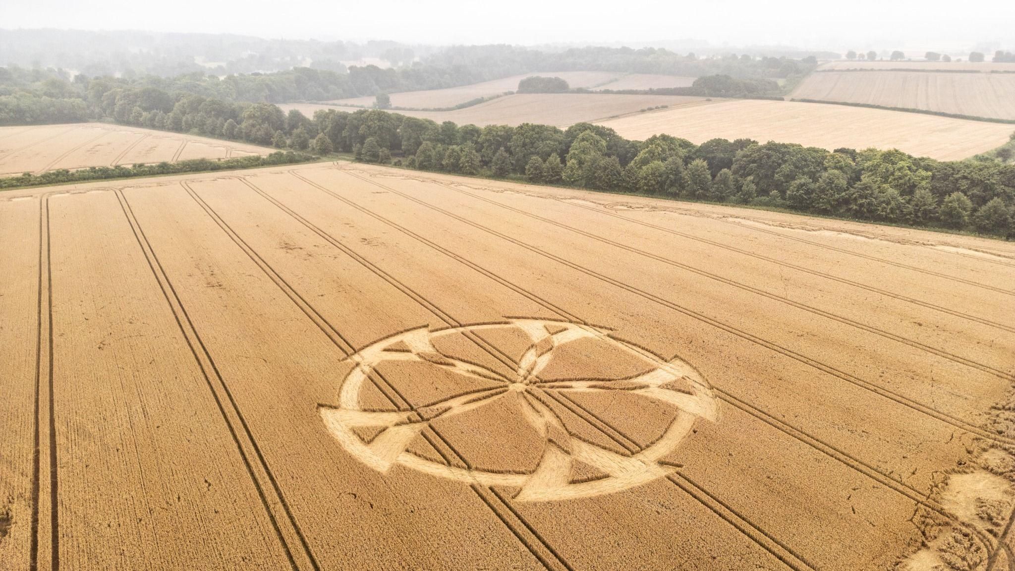 An aerial view of a crop circle resembling a five-spoked wheel with a rural landscape in the background