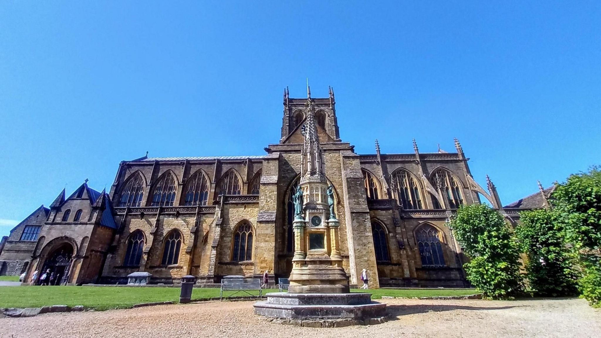 An abbey dominates the image with clear blue skies behind it in the foreground is a monument surrounded by gravel paving