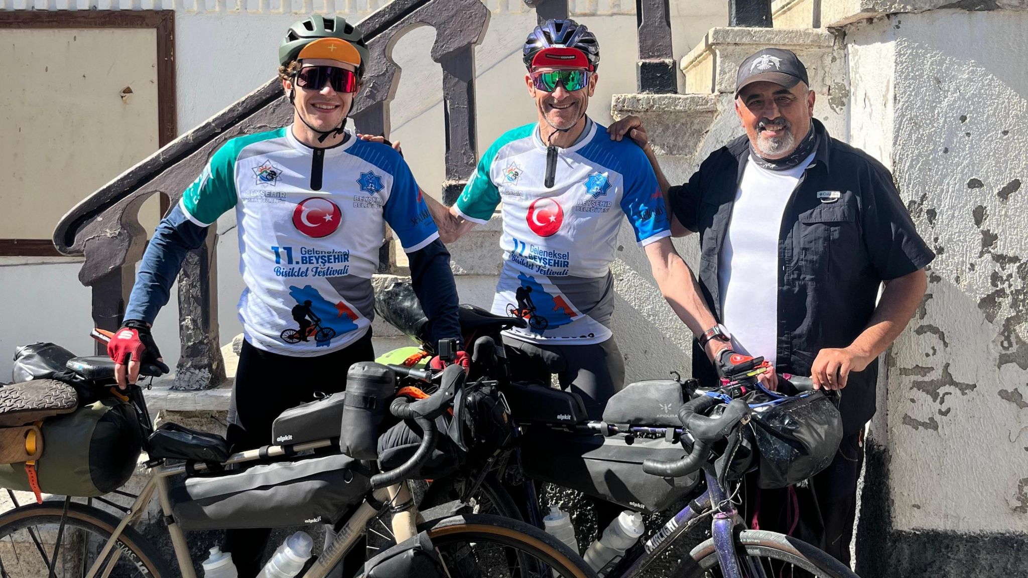 The cyclists in special white and blue cycling vests with a turkish flag symbol on them they are stood with their bikes, wearing sunglasses and stood next to a man who is wearing a hat