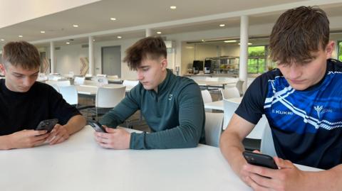 Three male pupils looking at their phones waiting for exam results