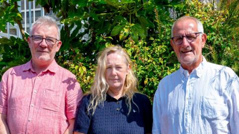 A man with glasses in a pink shirt, a woman with blonde hair in a blue top and a man with glasses in a light blue shirt.