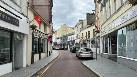 Ramsey high street, with two rows of shops including an opticians, a tattoo parlour and a pharmacy. Red Manx flags are dotted along the street above the shops. The tarmac of the road separating the rows is red, and cars are parked along the right-hand side of the street.