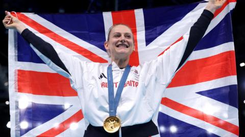 Bryony Page celebrating her gold medal win. She is in a GB tracksuit and holding a union jack over her shoulders. The gold medal is around her neck