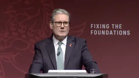 PM Sir Keir Starmer wearing a suit, making a speech at a lectern, standing in front of a backdrop with 'fixing the foundations' written on it 