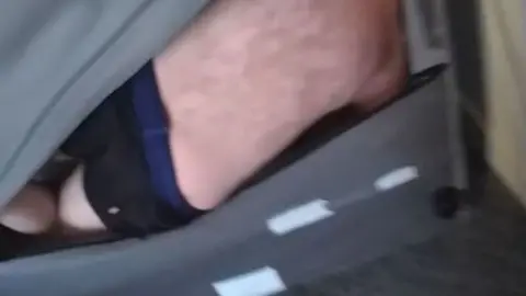 A blurry police-cam image of a man's pale back and dark pants. He is tucked into the back of a sofa with his knees raised. The grey fabric is parted to show his back