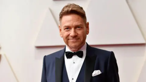 A man with short strawberry blonde hair and stubble, wearing a dark blue tuxedo with a black bow tie, smiling at the camera with a white wall behind him