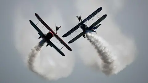 Two planes flying in the sky with smoke trails coming off the back. On the top of each plane is an acrobat.