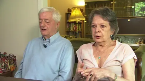 A man in a pale blue shirt and jumper alongside a woman in a pale pink blouse sitting at a dinning table in their home, with a lamp and a glass cabinet behind them