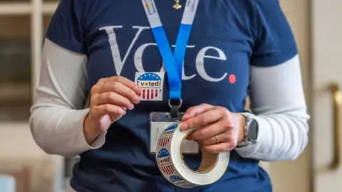 A US poll worker wearing a VOTE T-shirt holds a roll of "I voted!" stickers