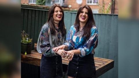 Georgia and Melissa with a garden fence behind them. They are both wearing shirts and denim skirts.