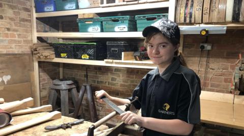 Sophie smiling into the camera as she works on a piece of wood in a workshop wearing an Adidas cap and Garners polo-shirt with a ponytail