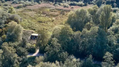 A drone photo taken looking down on a hut in the middle of a park with grass and trees 