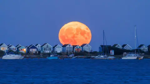 Jack Lodge Photography A large orange moon rises in a deep blue sky over colourful beach huts on Mudeford spit. The sky behind is clear some of the colouful beach huts windows are lit up from inside. There are several small boats floating in the foreground on a dark blue sea.