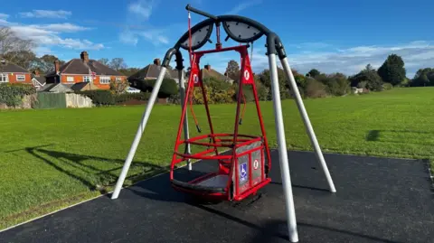 A red, wheelchair-accessible swing in a park. It is on a black base and surrounded by a green grass field.