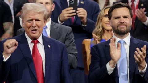 Trump wearing red tie with white plaster bandage on his ear, he is clasping his fist and smiling beside JD Vance in a blue tie clapping and slightly smiling